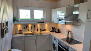 A compact kitchen with a sink, stove, and oven, wooden cabinets, and a window above the sink. - Schwalbennest - Infrared Sauna, Fireplace, 4 Sun Loungers, EV Charging Station (Olpenitz)