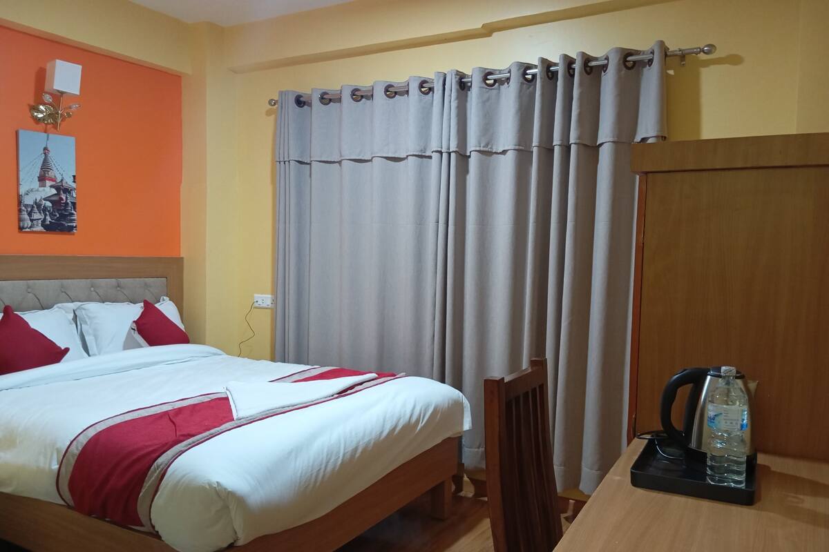 A hotel room with a bed, a wooden chair, a table with a kettle and water bottle, and a curtain.