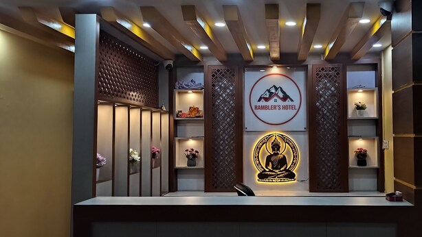 Uma área de recepção com design moderno, apresentando um balcão de atendimento, uma estátua de Buda e uma placa indicando o Hotel Ramble's.