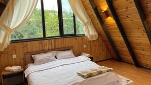 Een slaapkamer met een houten plafond, een groot bed, wit beddengoed en uitzicht op groen door de ramen.