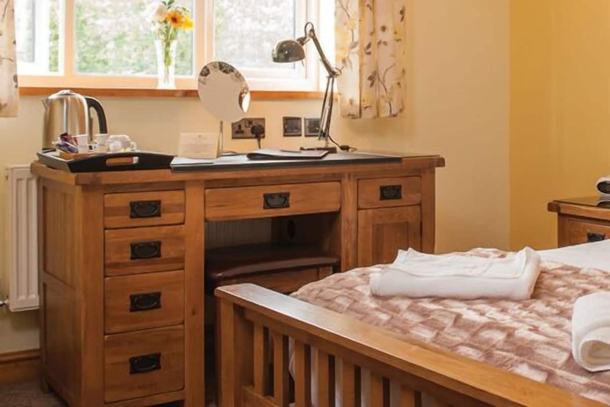 A neatly arranged bedroom with a wooden bed, a bedside table with a lamp, a nightstand with a phone, and a window with curtains.