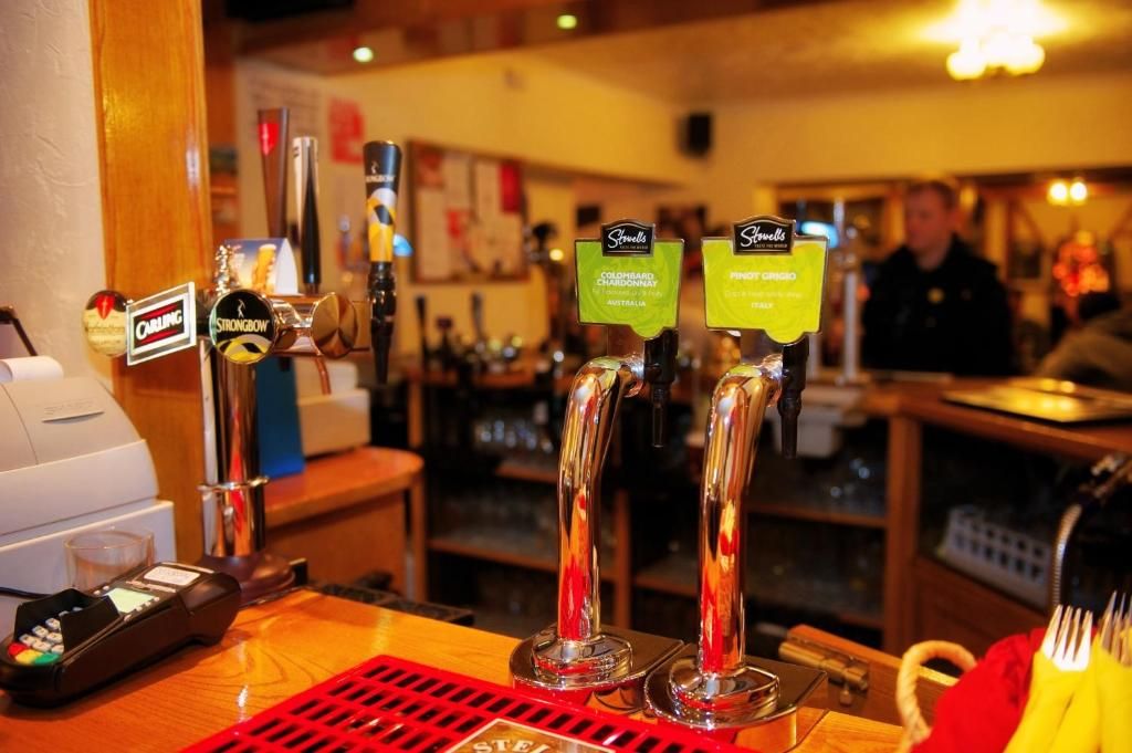 Zona de bar amb diverses aixetes etiquetades amb marques de cervesa i un terminal de pagament.