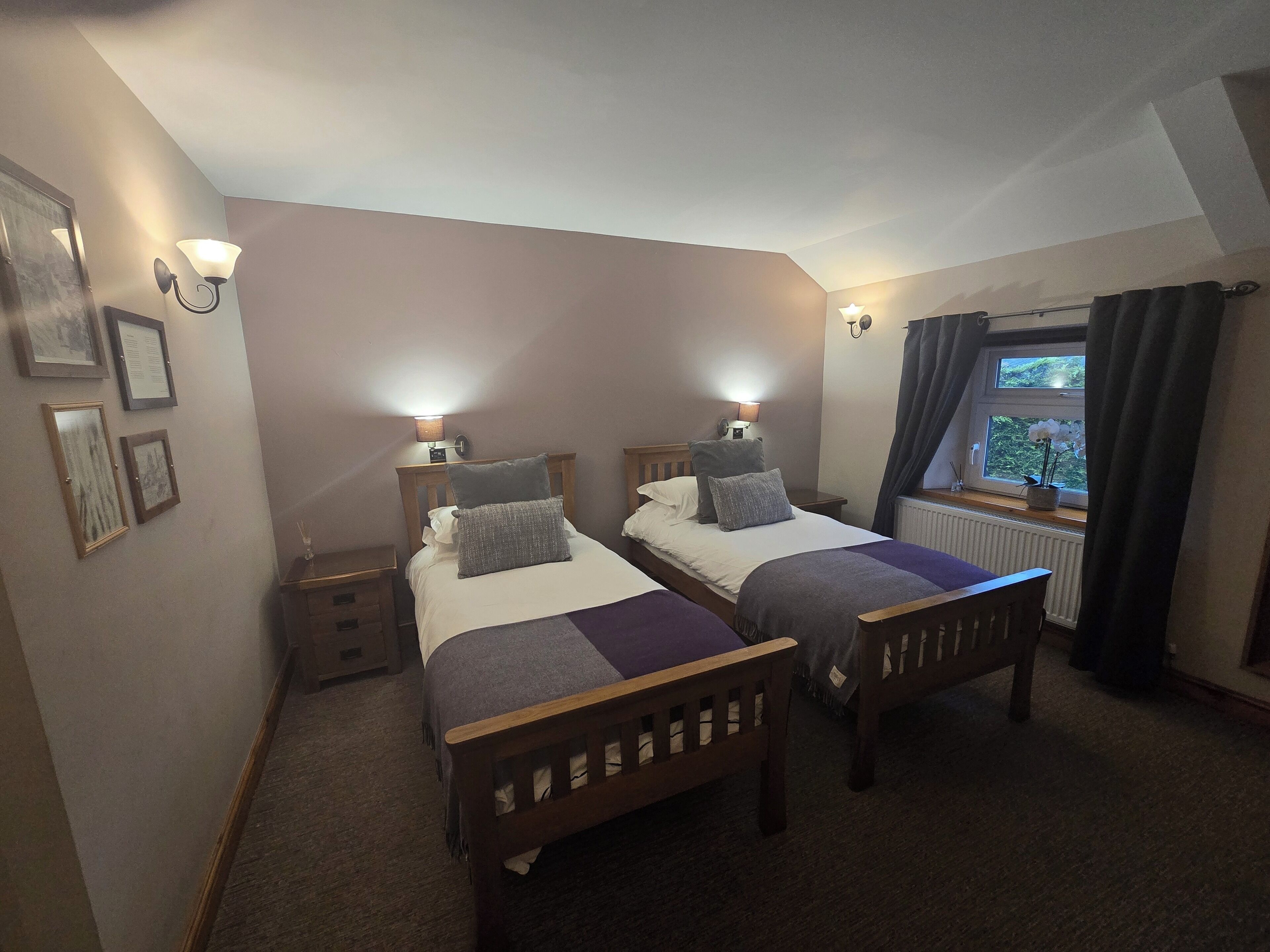 A bedroom with two single beds, bedside tables, wall-mounted lamps, framed pictures, and a window with curtains.