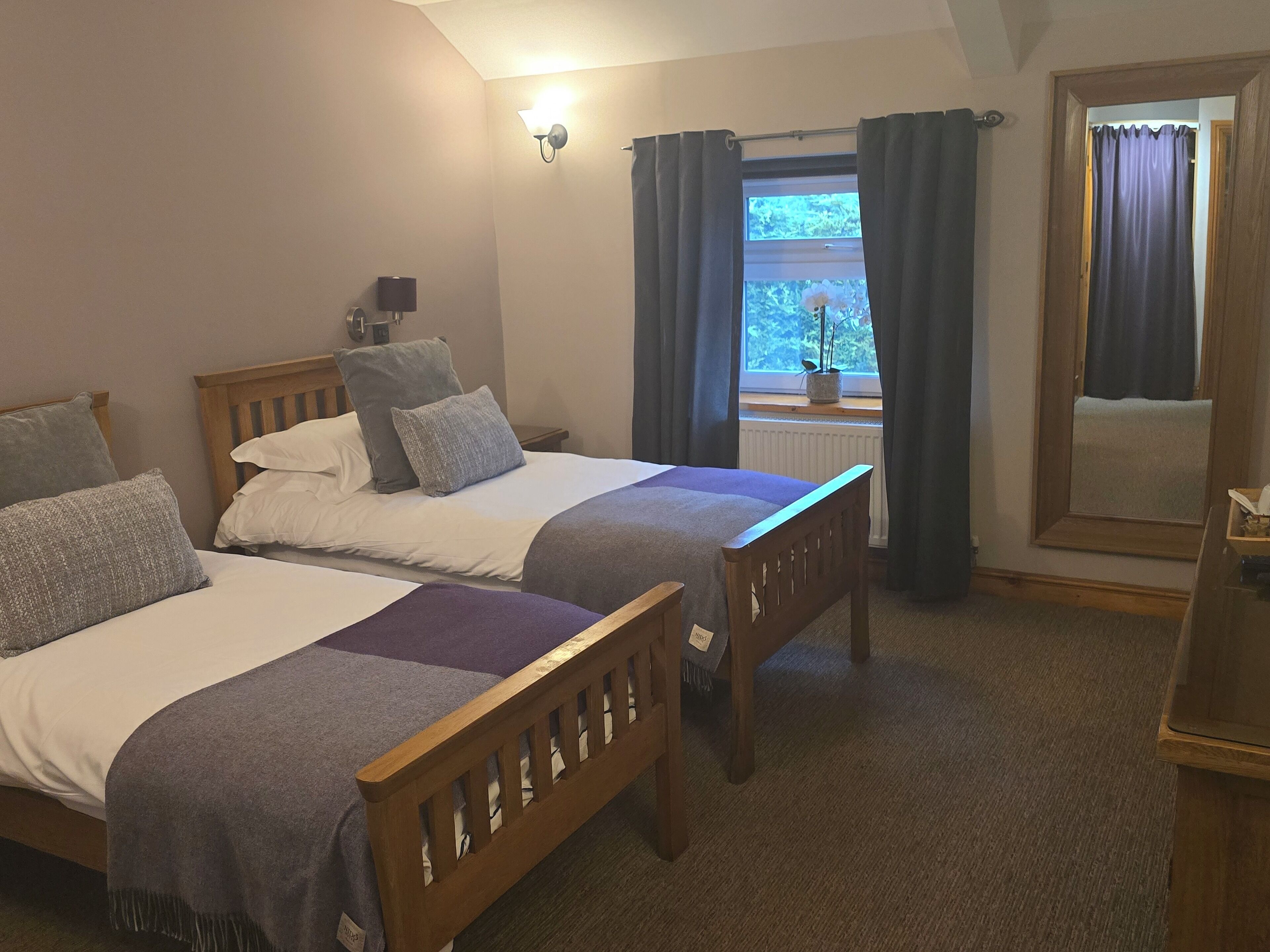 A bedroom with two wooden beds, a nightstand, a lamp, a window with curtains, and a mirror.