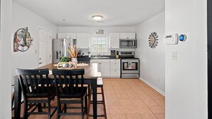 A modern kitchen with white cabinets, a stainless steel refrigerator, a microwave, and a built-in oven. There is a dining table with chairs and a decorative wall piece.