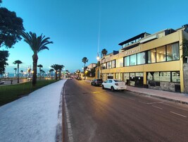 Um hotel com fachada moderna, situado numa rua costeira com palmeiras e vista para o mar.