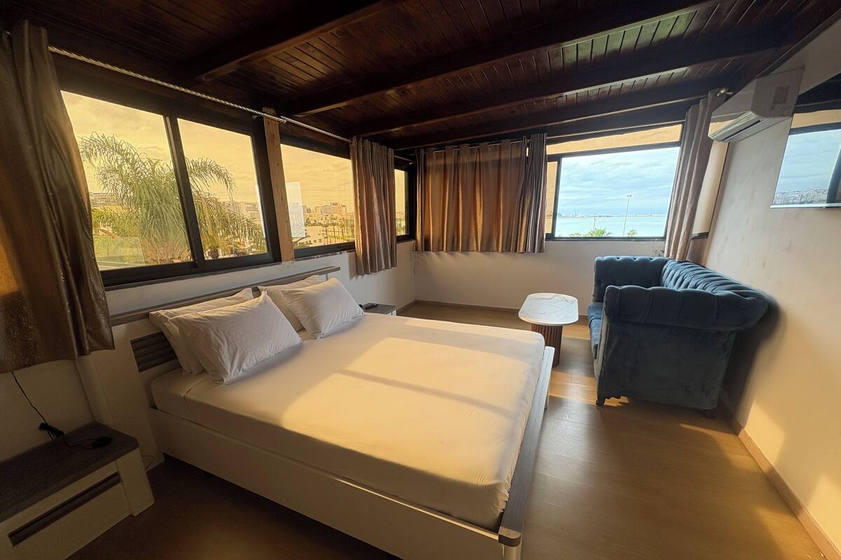 A room with a bed, a small table, a chair, and large windows offering a view of the city and water.