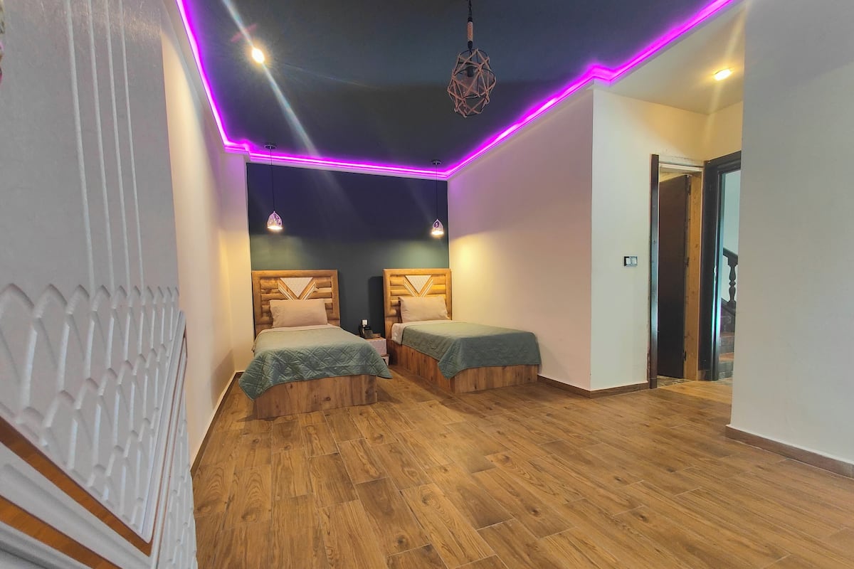 A room with two wooden beds, a patterned wall, and a ceiling with purple LED lighting.