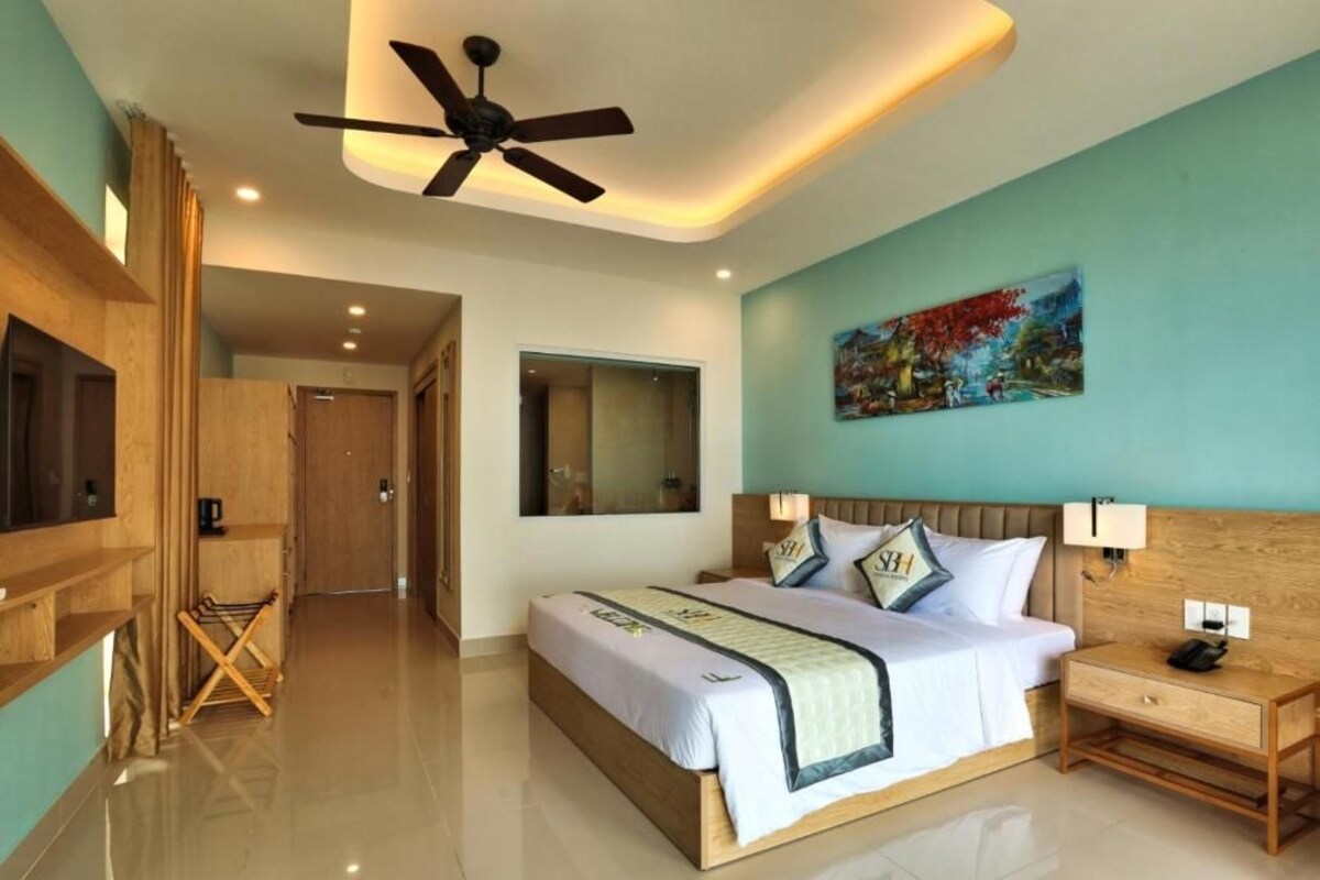 A modern hotel room with a large bed, a ceiling fan, a painting on the wall, and a wooden nightstand.