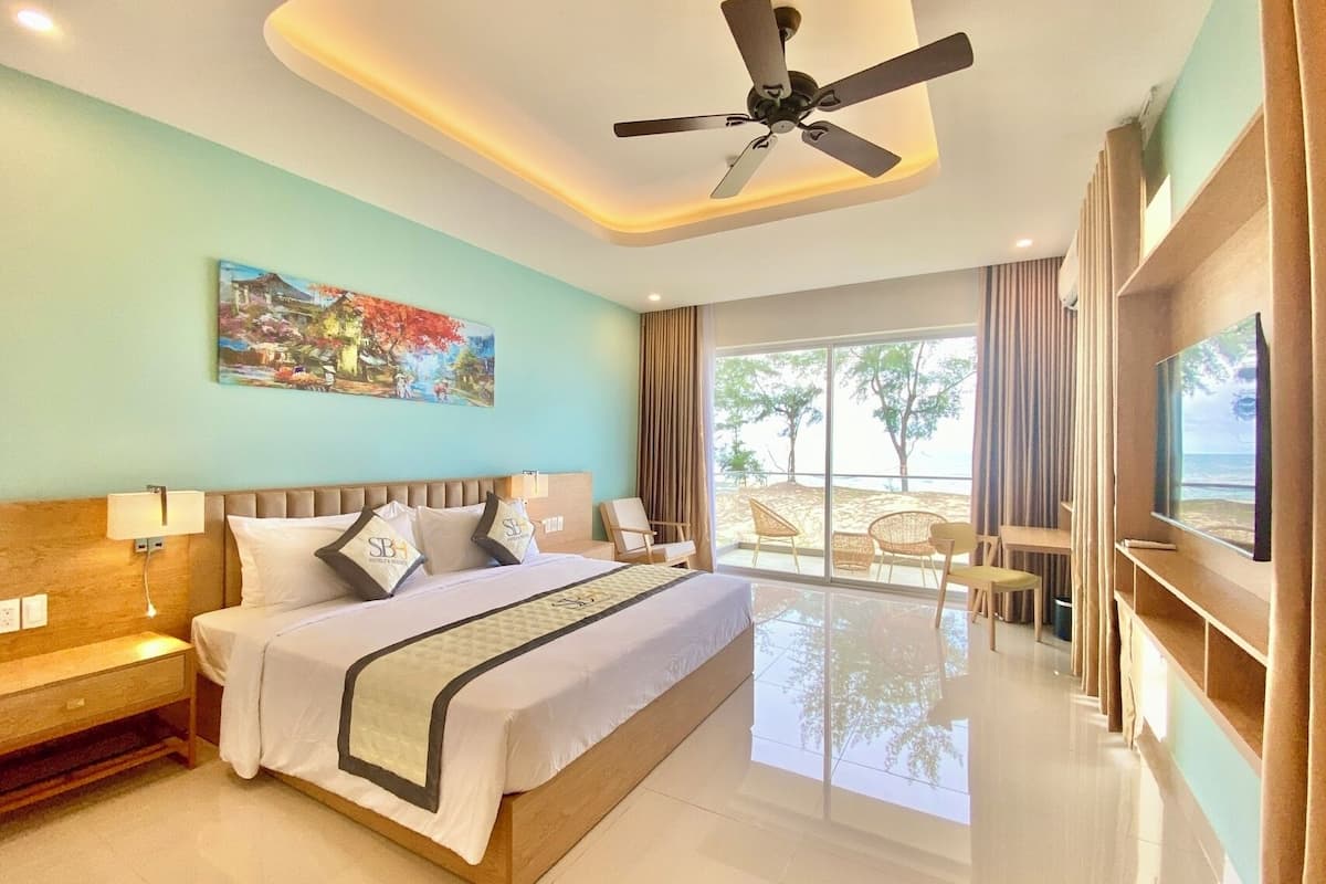 Habitación de hotel con cama, ventilador de techo, un cuadro y vistas a la playa.