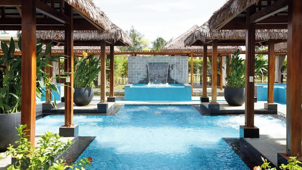 A pool area with a central water feature, surrounded by wooden structures with thatched roofs, and potted plants.