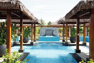 Un área de piscina con una fuente central, rodeada de estructuras de madera con techos de paja y plantas en macetas.