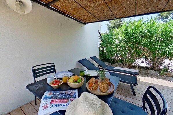 An outdoor dining area with a table set for two, featuring a newspaper, fruits, and a pineapple.