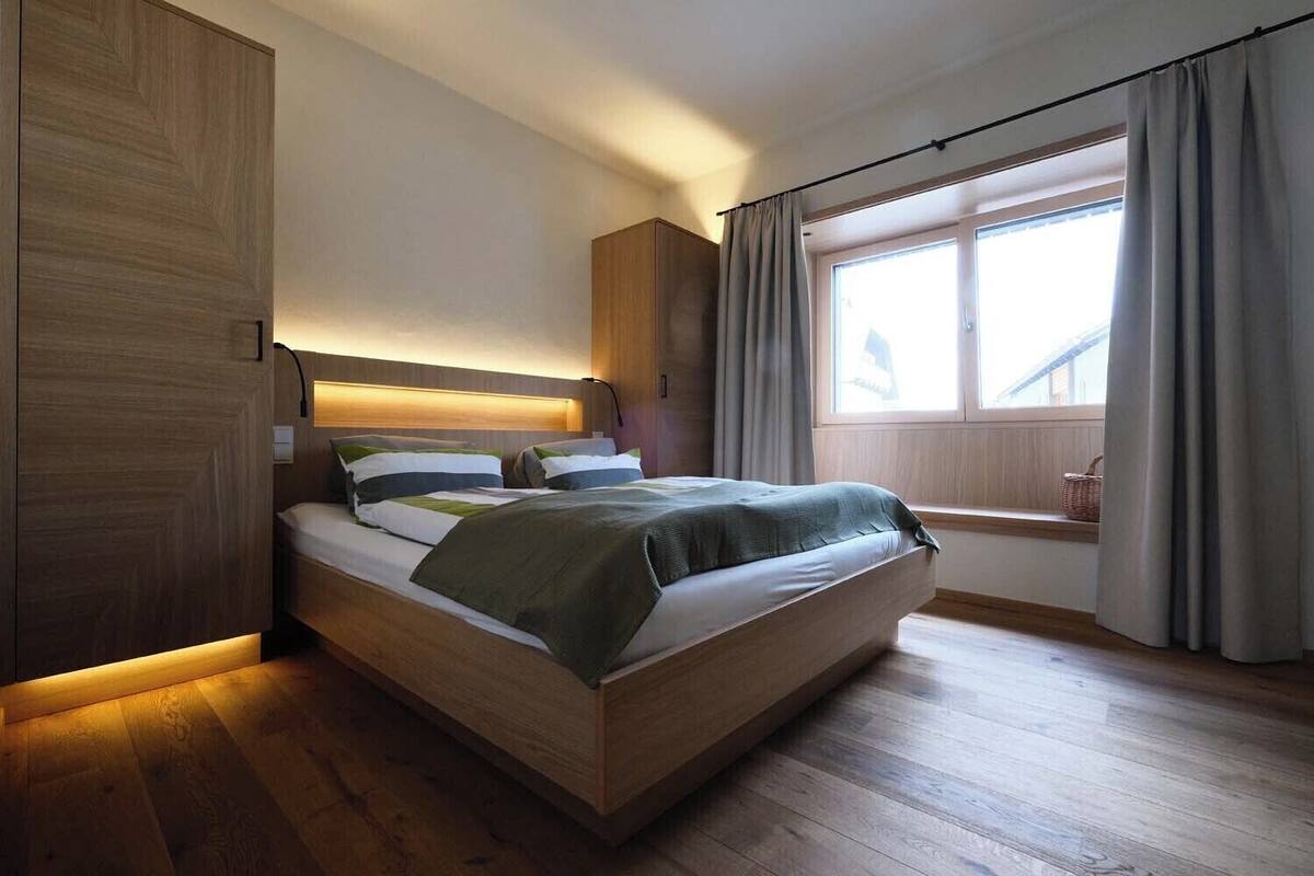 Un dormitorio moderno con cama de madera, cabecero con iluminación integrada, ventana con cortinas y suelo de madera.