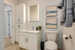 Une salle de bain moderne avec un lavabo blanc, des toilettes et un miroir. Des serviettes sont accrochées au mur et une poubelle se trouve à côté des toilettes.