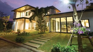 Una casa moderna de dos pisos con una entrada bien iluminada, una escalera visible y un jardín con un sendero que conduce a la puerta principal.