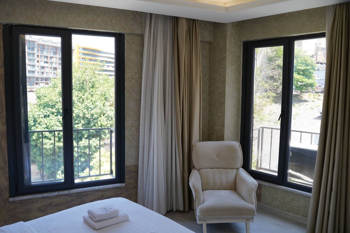Habitación de hotel con cama, una silla y amplias ventanas que ofrecen vistas a un edificio y vegetación.