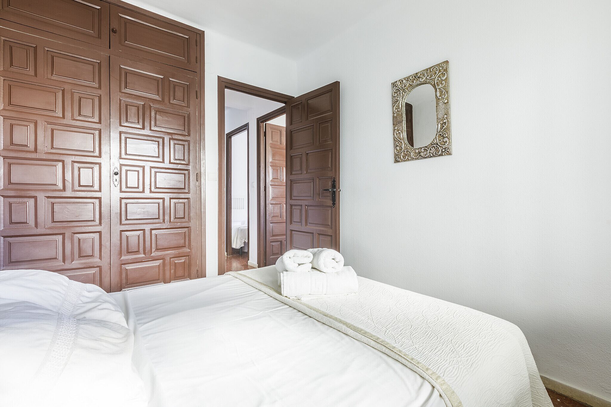 Habitación de hotel con un amplio armario de madera, una cama con ropa blanca y un espejo decorativo en la pared.