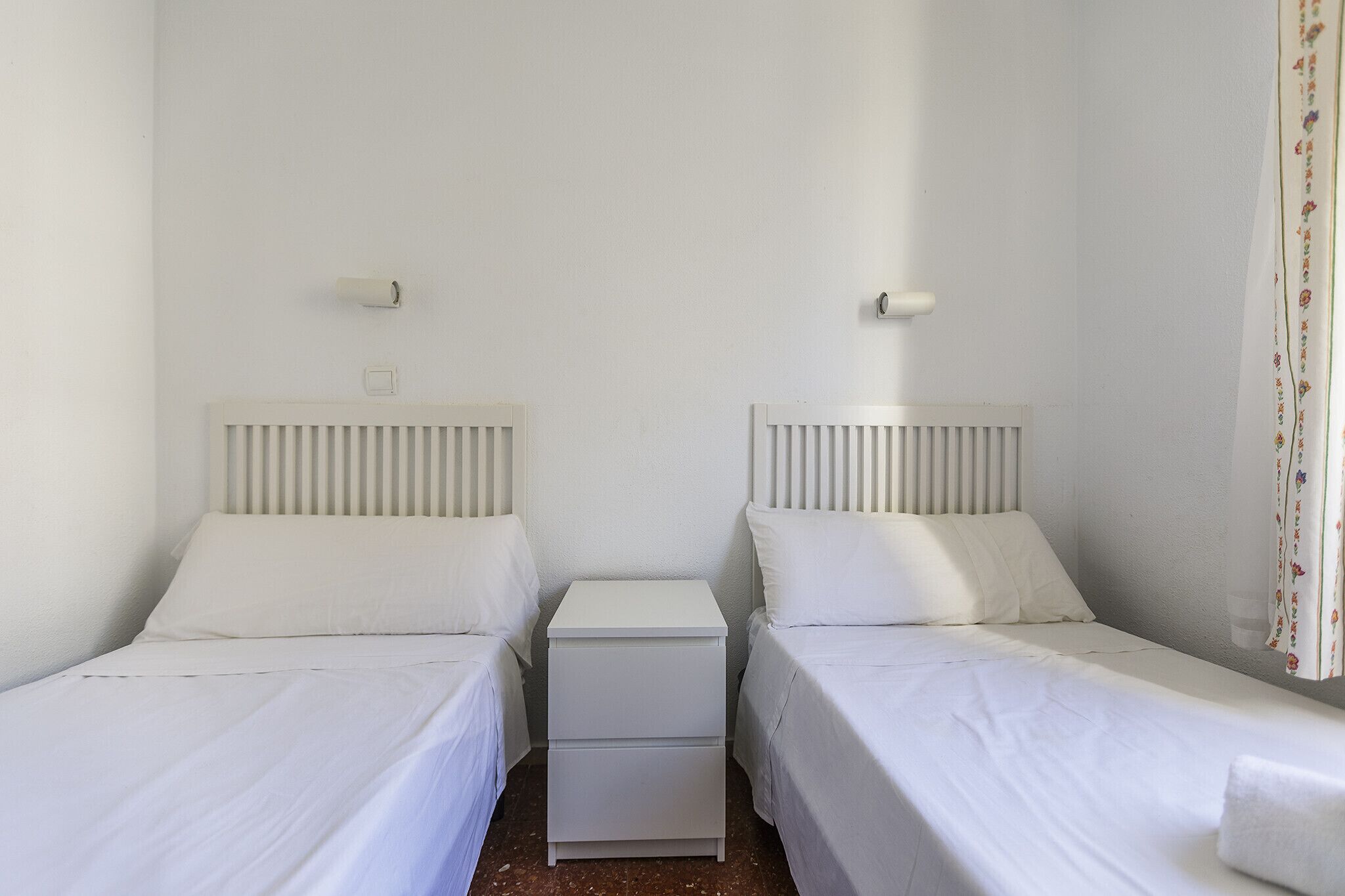 A simple, clean room with two single beds, a small bedside table, and a radiator.