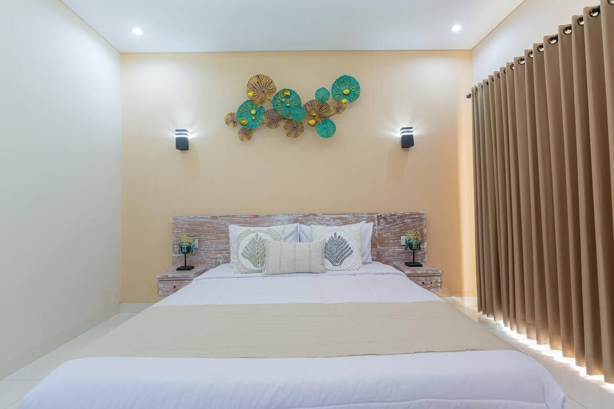 Un dormitorio moderno con una cama grande, dos mesitas de noche con lámparas, un adorno en la pared y una ventana con cortinas.