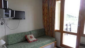 Una habitación con un sofá verde, una mesita, un televisor montado en la pared y una ventana con cortinas.