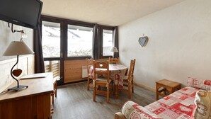 A cozy room with a wooden desk, a round table with chairs, a bed with a patterned blanket, and a large window with a view of a snowy landscape. - travelski home select - Vue Pistes - Studio cabine 3/4 personnes Etoile F16 (La Plagne)