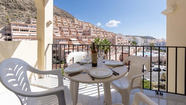 Un balcon avec une table dressée pour deux personnes, d’où l’on peut admirer une ville côtière, avec ses bâtiments et une montagne en arrière-plan.