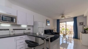 Une cuisine moderne avec des meubles blancs, un four à micro-ondes et un réfrigérateur. On y trouve un tabouret de bar noir et une table en verre. Dans l’espace de vie, il y a un canapé noir et une table à manger avec des chaises. Un balcon avec des rideaux bleus et une vue sur les immeubles est visible.