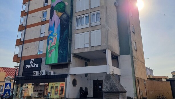 A multi-story building with a large mural on the side, a storefront with an 'optika' sign, and a bicycle parked outside.