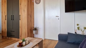 A living room with a wooden wardrobe, a white door, a flat-screen TV, a grey sofa, and a small table with decorative items.