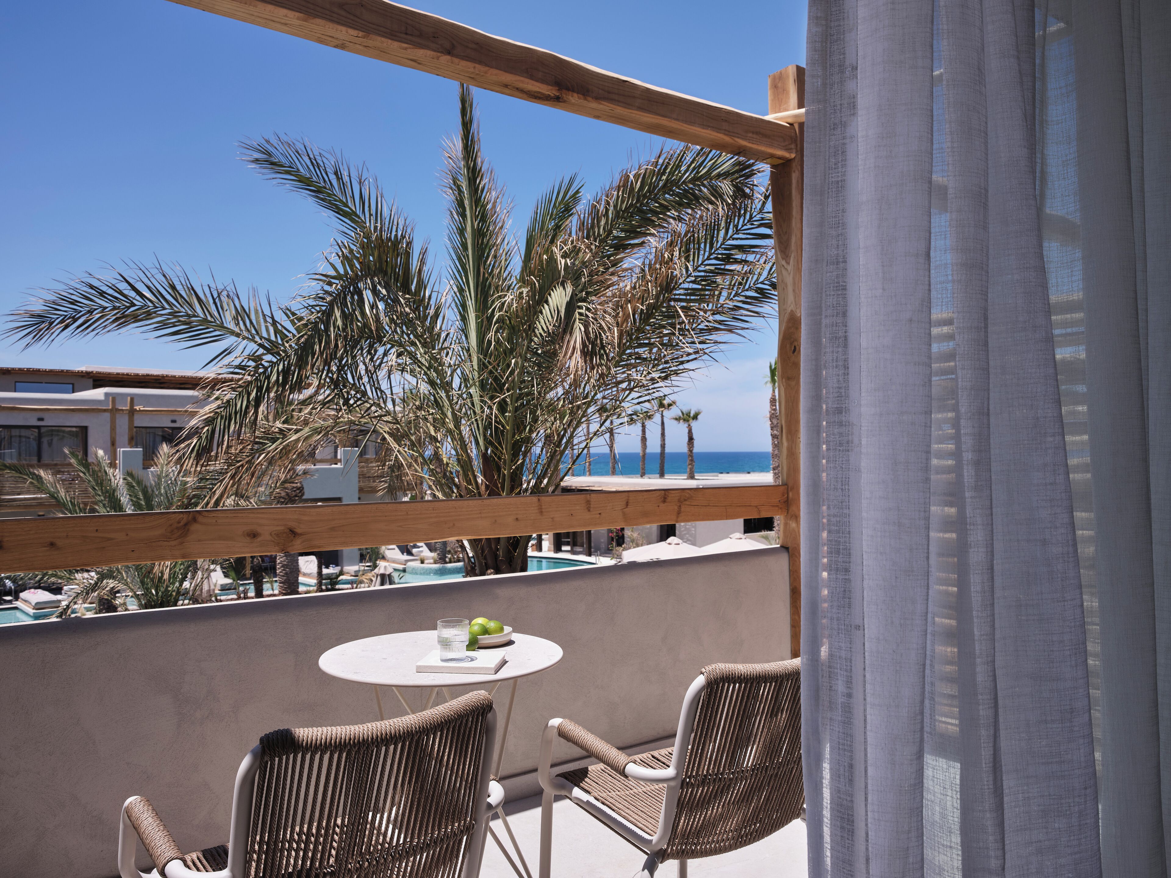 A balcony with wicker chairs, a small round table with a glass of water and a book, and a view of the sea and palm trees.
