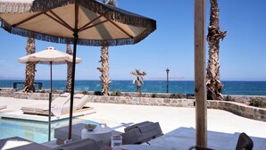 Zona junto a la piscina con sillas de descanso, una mesa y sombrillas, con vista al mar azul y cristalino.