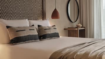 Una cama bien hecha con dos almohadas, una lámpara colgante, un espejo redondo y una mesita de madera con un jarrón.