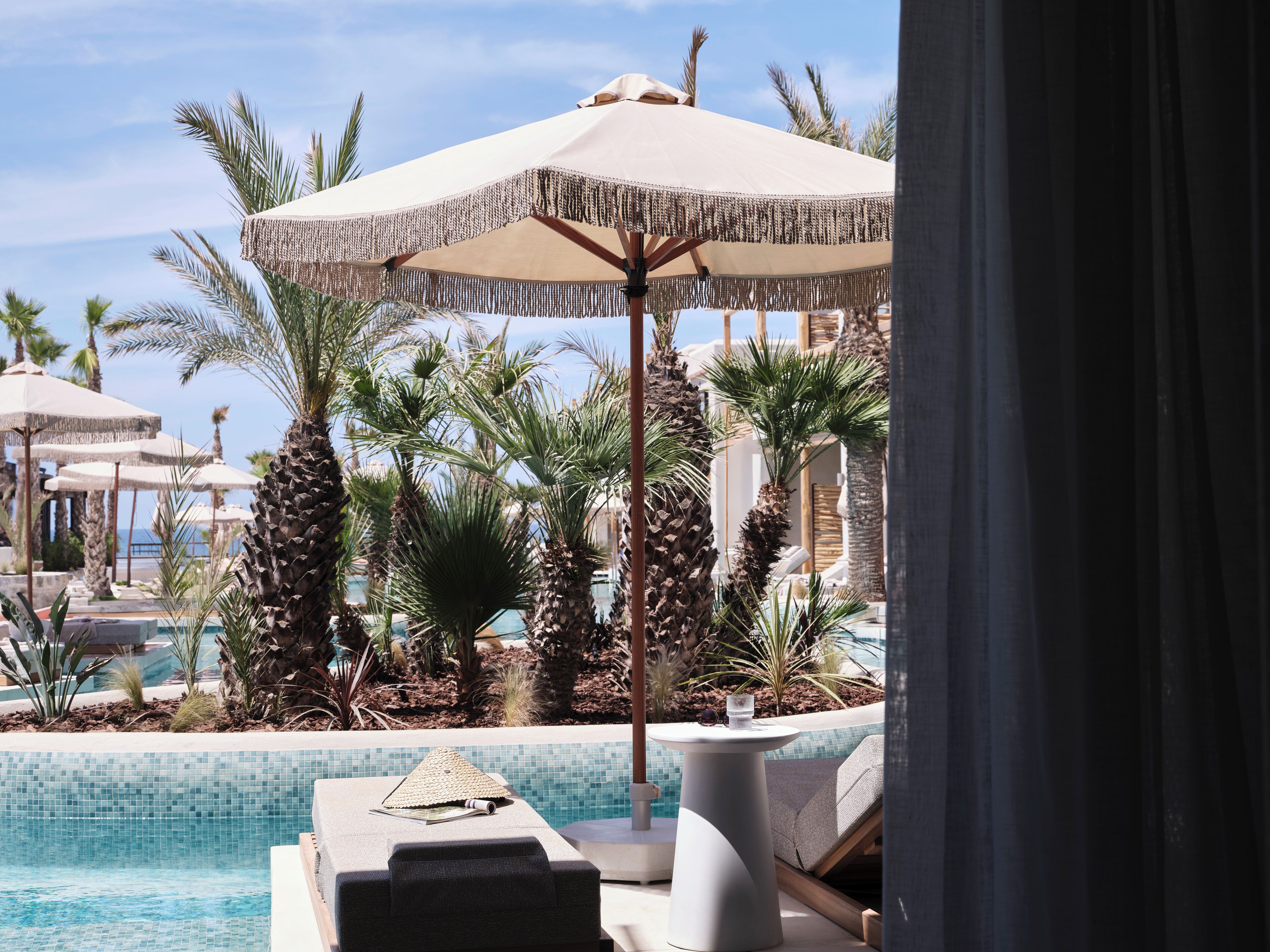 A poolside area with lounge chairs, a covered umbrella, and palm trees.
