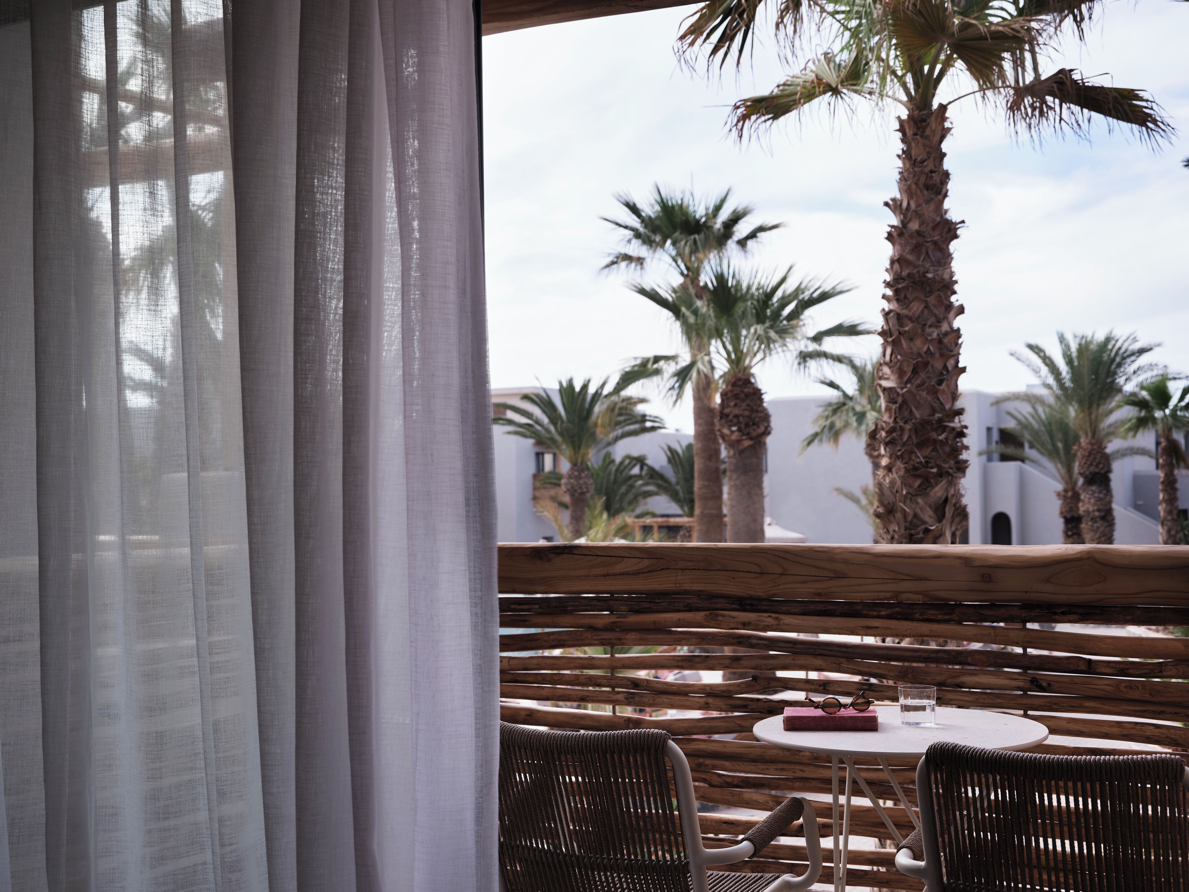 A terrace with wicker furniture, a table with a book and glasses, and palm trees in the background.