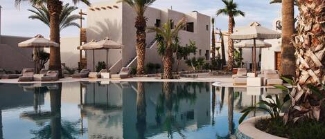 A pool area with lounge chairs and umbrellas, surrounded by palm trees and a building with a modern design.