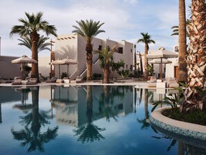 Zona de piscina con sillas de descanso y sombrillas, rodeada de palmeras y un edificio de diseño moderno.