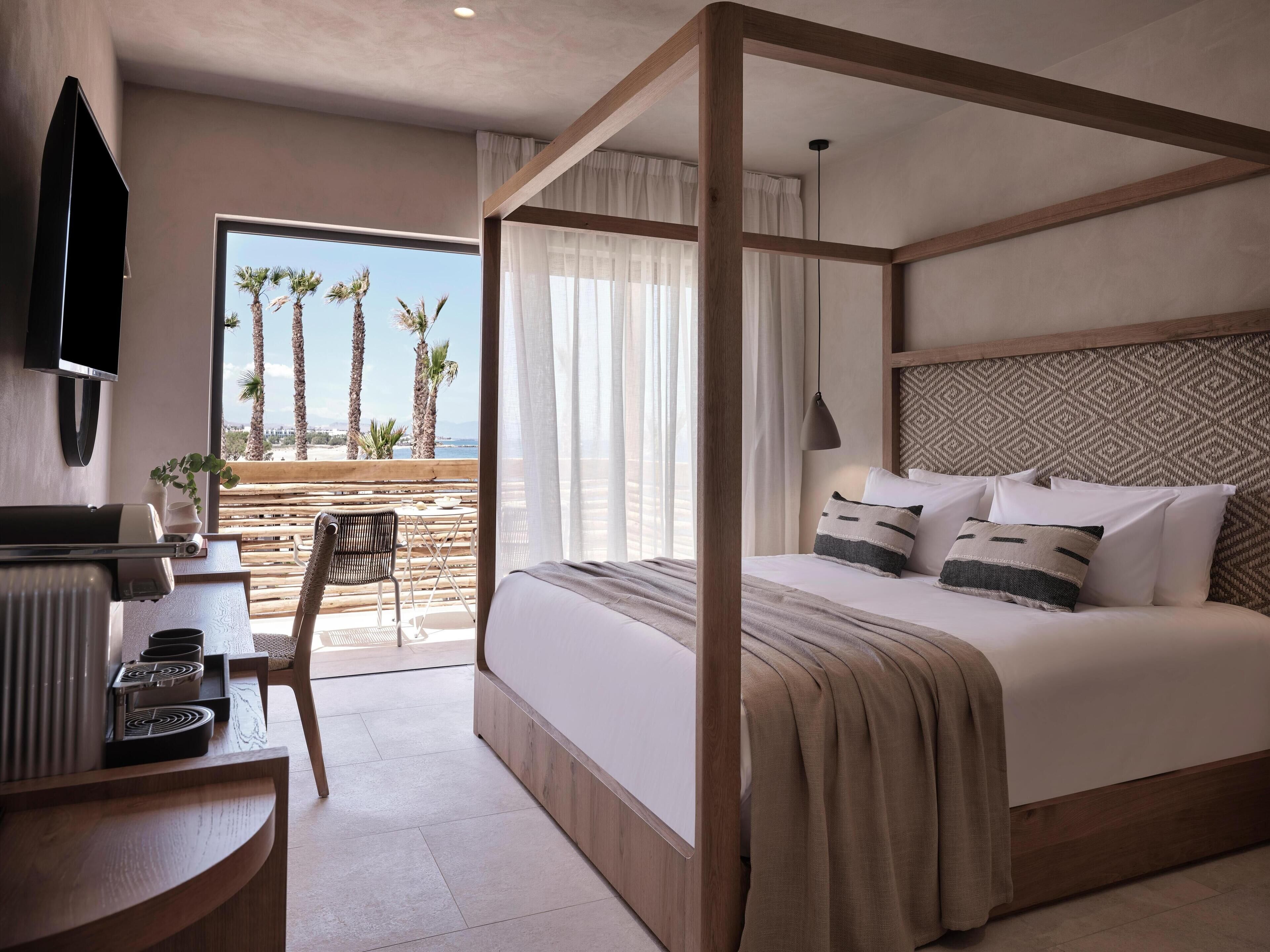 Un dormitorio con cama con dosel, un escritorio con televisor de pantalla plana y vista a palmeras y la playa.