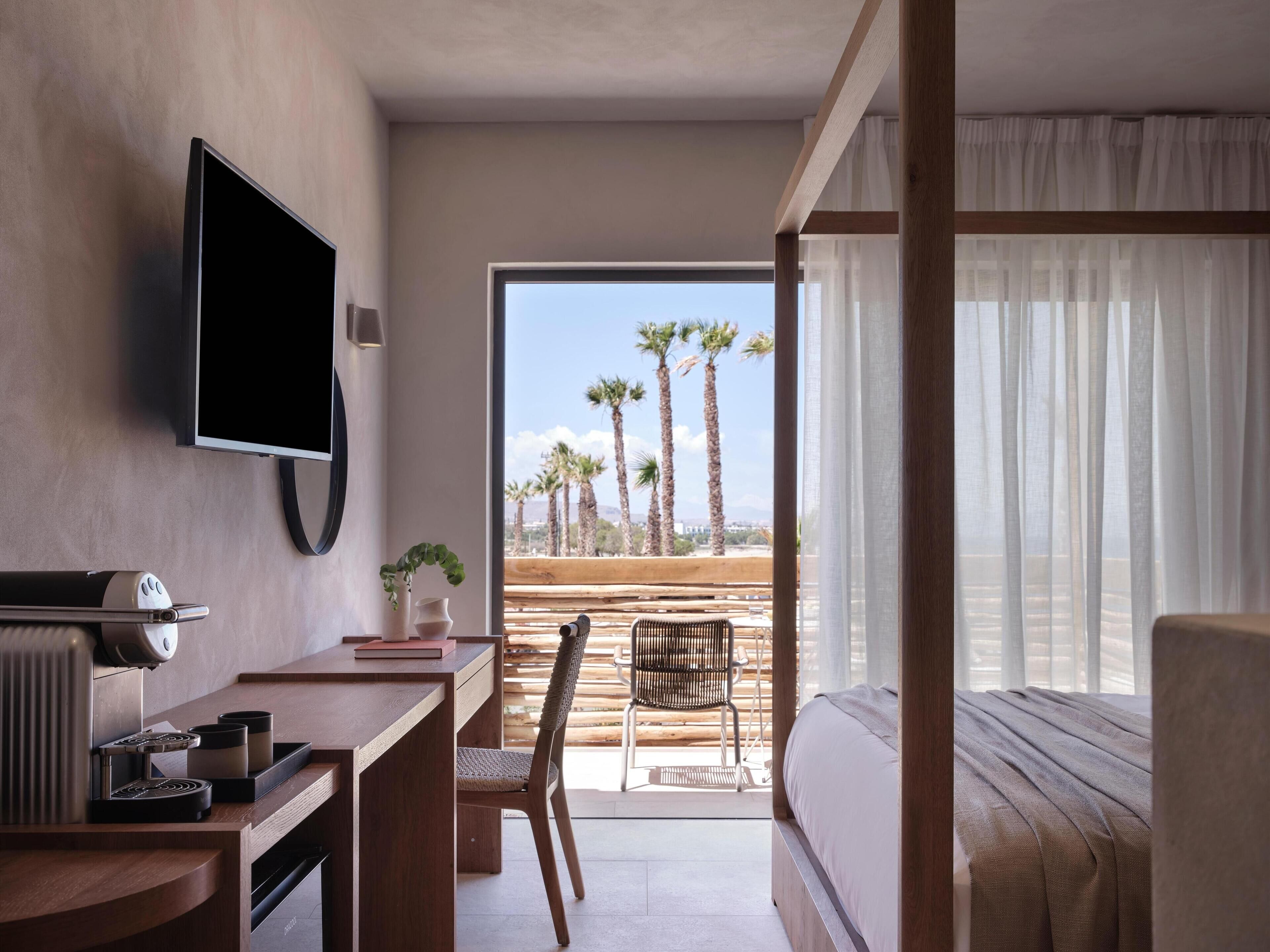 A modern hotel room with a bed, a desk with a coffee machine, a TV, and a view of palm trees.