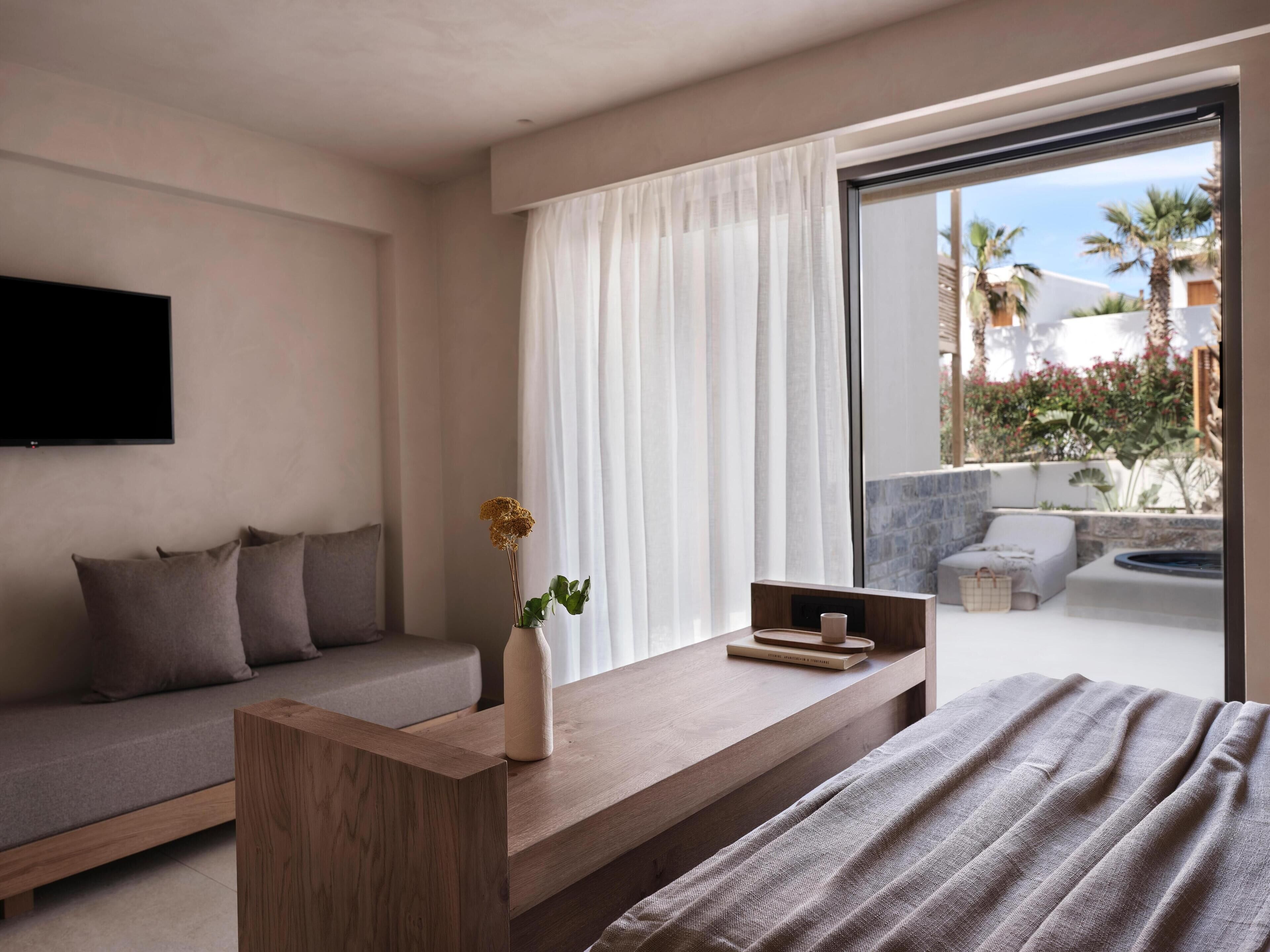Una habitación de hotel moderna con sofá, una mesita de madera, un ventanal con cortinas y vistas al exterior.