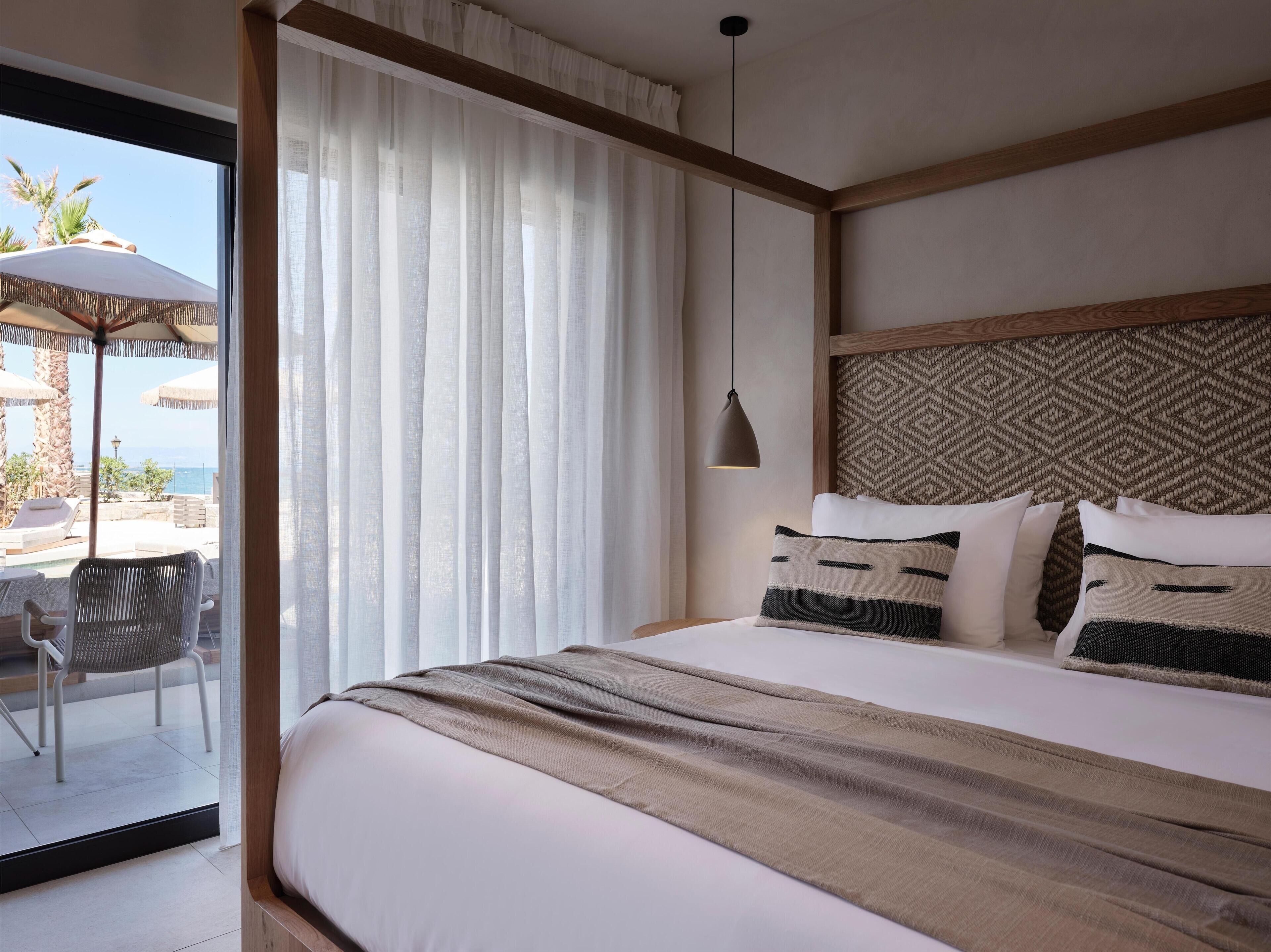 Habitació d'hotel amb un llit gran, vista al mar a través de cortines translúcides, un panell de capçal teixit i una làmpada penjant.