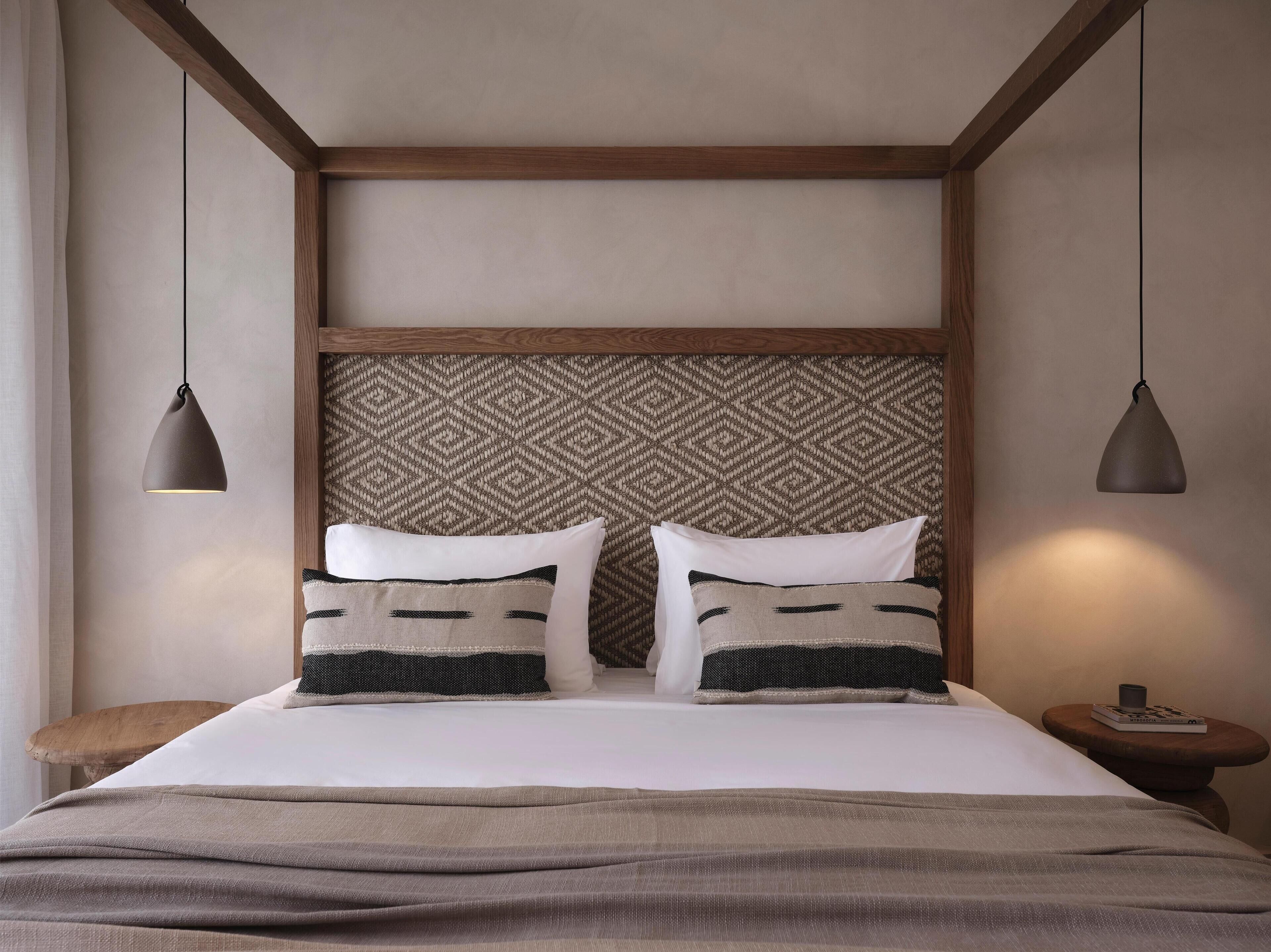 A bedroom with a wooden headboard, two bedside tables, a bed with white bedding and patterned pillows, and pendant lights.
