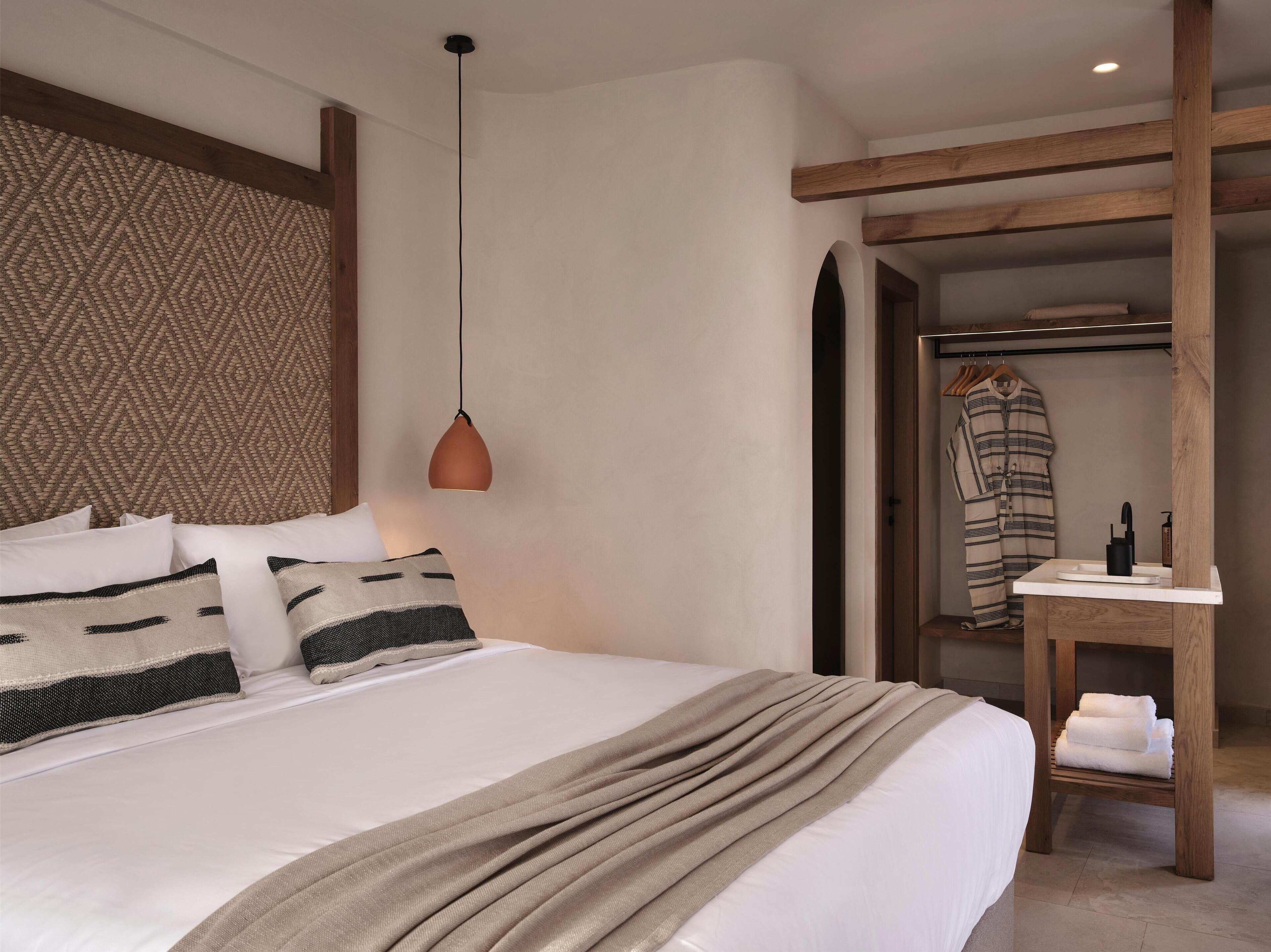 A neatly made bed with a patterned headboard, a hanging pendant light, and a wooden bench with towels.