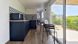 A modern kitchen with a black countertop, a microwave, and a patterned backsplash. There is a dining area with a table and chairs, and a view of greenery outside. - 2. Turquoise - HOME CASSIS - Maison Méditerranée (CASSIS)