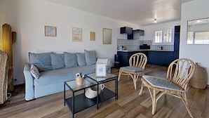 Uma sala de estar moderna com um sofá cinzento, uma mesa de centro de vidro, cadeiras de vime e uma área de cozinha ao fundo.