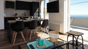 A modern kitchen with black cabinets, a dining area with four chairs, a TV mounted on the wall, and a balcony with a view of the sea. - 2. Indigo - HOME CASSIS - Maison Méditerranée (CASSIS)