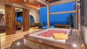 A luxurious spa area with a large bathtub filled with rose petals, wooden flooring, and a view of the city at dusk.
