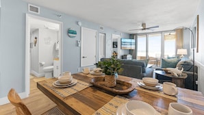 Un comedor con una mesa de madera preparada para cuatro personas, una planta en maceta y un televisor al fondo.