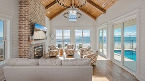 A spacious living room with a fireplace, large windows, and a view of the ocean. - Oceanfront Beach House, Elevator to First Floor, Ocean View Pool, Dog Friendly (Pawleys Island)