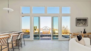 A spacious living area with large windows, a bar area with stools, and a view of a desert landscape. - The Tree House By The Cohost Company (Joshua Tree)